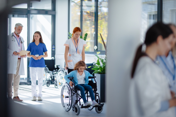 Young nurse pushing little girl on wheelchair at a hospital corridor.