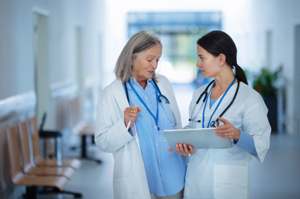 Older doctor giving advise to her younger colleague, discussing at a hospital corridor. Health care concept.