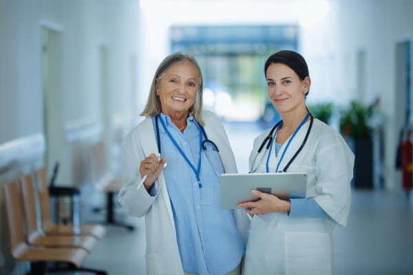 Older woman doctor giving advise to his younger colleague, discussing at a hospital corridor. Health care concept.