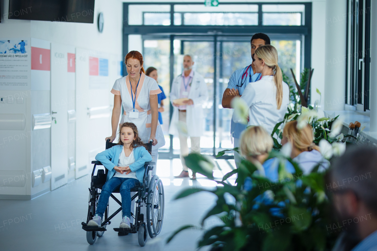 Young nurse pushing little girl on wheelchair at a hospital corridor.