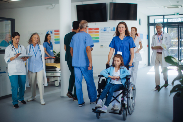 Young nurse pushing little girl on wheelchair at a hospital corridor.