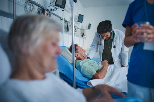 Young nurse giving glass of water to senior patient, doctor laughing with patient at a hospital ward.