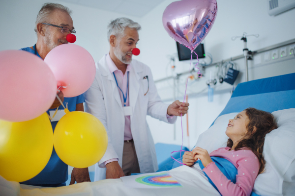 Happy doctors with clown red noses celebrating birthday with little girl in a hospital room.