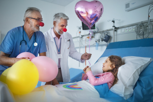 Happy doctors with clown red noses celebrating birthday with little girl in a hospital room.