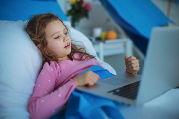 Little sick girl lying alone at hospital bed and using a laptop.