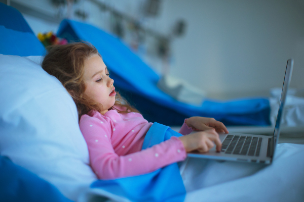 Little sick girl lying alone at hospital bed and using a laptop.