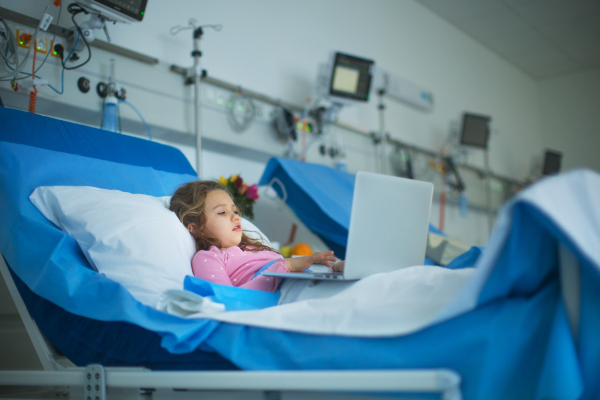 Little sick girl lying alone at hospital bed and using a laptop.