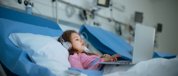 Little sick girl lying alone at hospital bed and using a laptop.