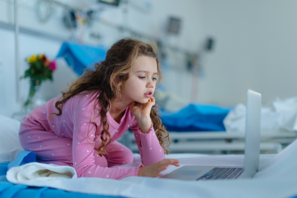 Little sick girl lying alone at hospital bed and using a laptop.