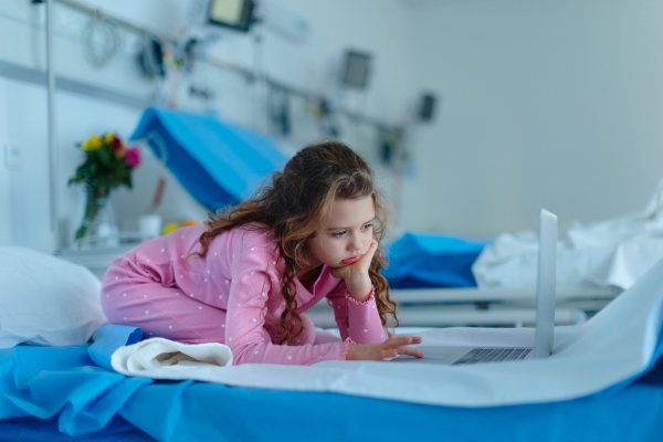 Little sick girl lying alone at hospital bed and using a laptop.