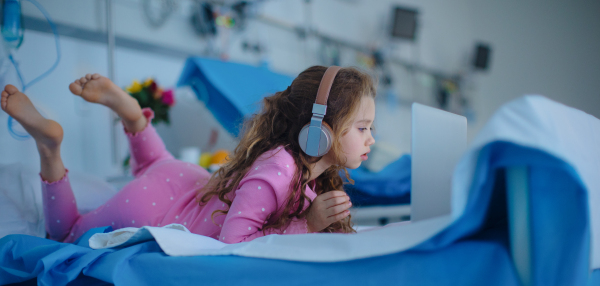 Little sick girl lying alone at hospital bed and using a laptop.