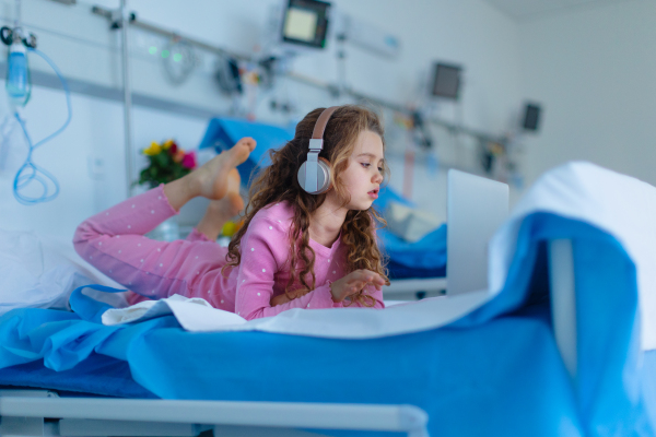 Little sick girl lying alone at hospital bed and using a laptop.