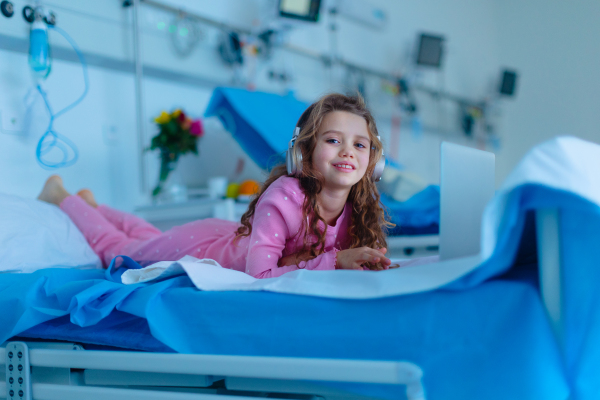 Little sick girl lying alone at hospital bed and using a laptop.