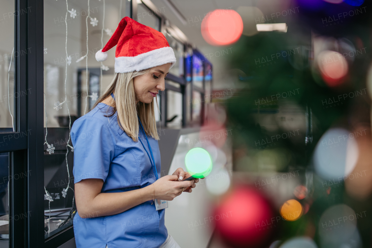 Portrait of beautiful nurse with christmas hat in hospital corridor, holding smartphone. Working Working in hospital on the Christmas day, Christmas Eve. Female doctor working a Christmas shift and can't be with her family.