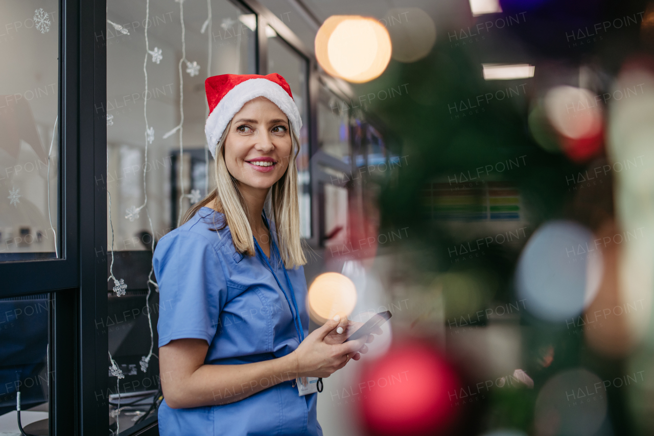 Portrait of beautiful nurse with christmas hat in hospital corridor, holding smartphone. Working Working in hospital on the Christmas day, Christmas Eve. Female doctor working a Christmas shift and can't be with her family.