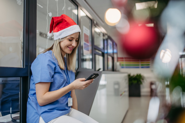 Portrait of beautiful nurse with christmas hat in hospital corridor, holding smartphone, reading text message. Working Working in hospital on the Christmas day, Christmas Eve. Female doctor working a Christmas shift and can't be with her family.