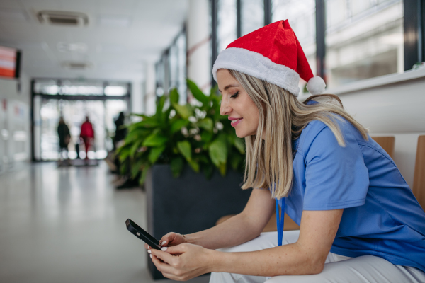 Beautiful nurse with christmas hat in hospital waiting area, holding smartphone, reading text message. Working Working in hospital on the Christmas day, Christmas Eve. Female doctor working a Christmas shift and can't be with her family.