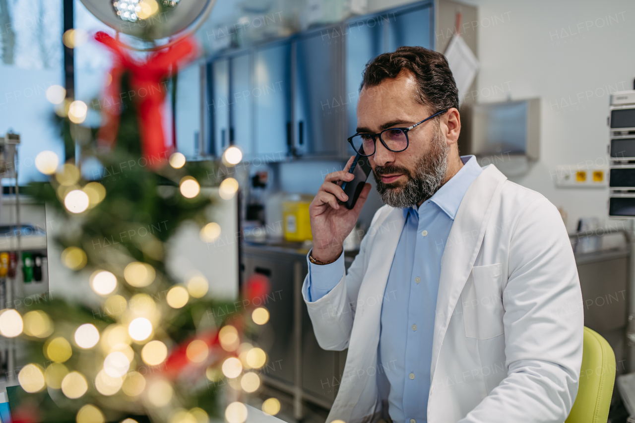 Portrait of busy doctor phone calling in emergency room, consulting room decorated for Christmas. Mature male doctor working a Christmas shift in the hospital, can't be with his family during Christmas Day and Christmas Eve.