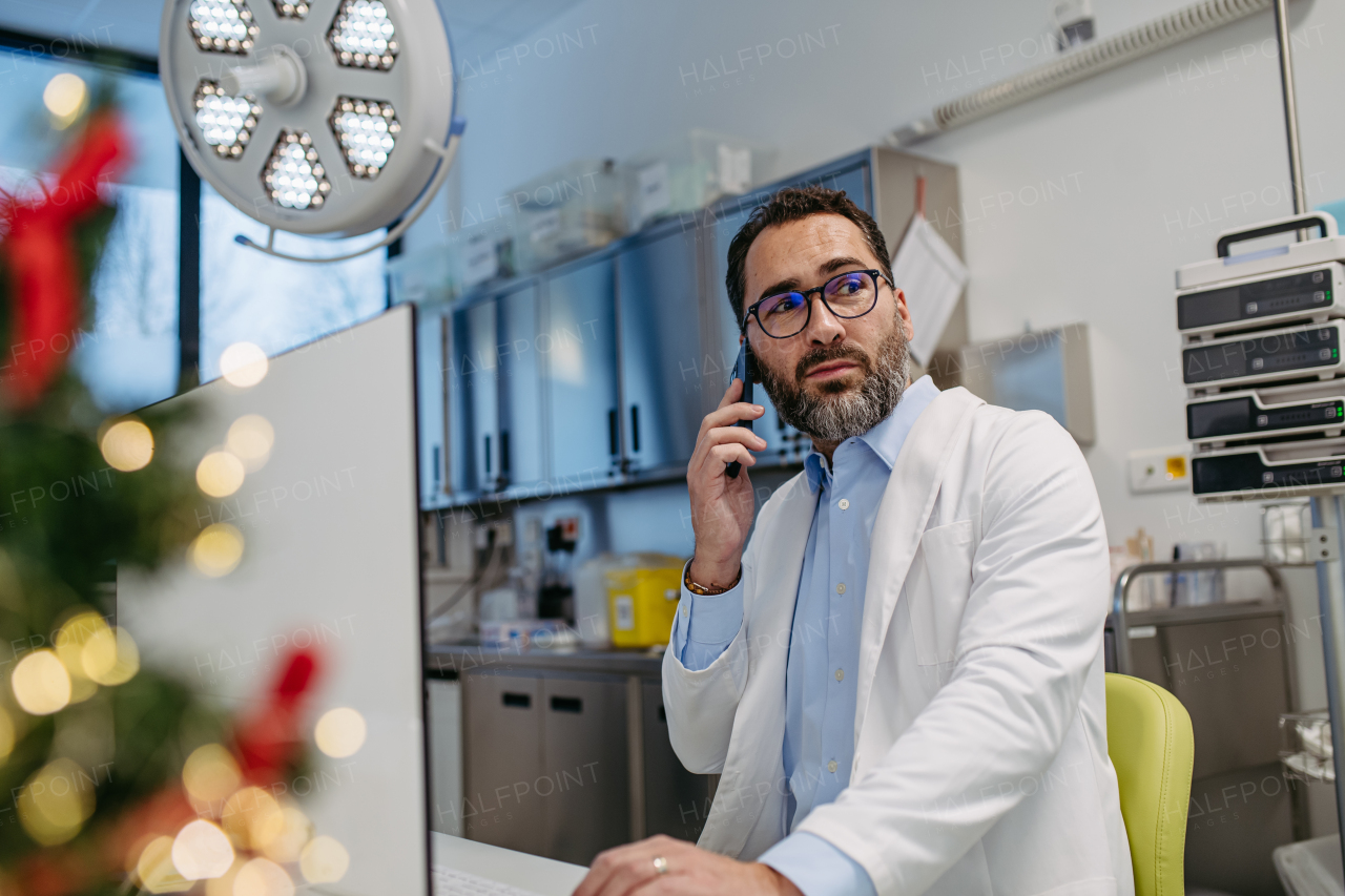 Portrait of busy doctor phone calling in emergency room, consulting room decorated for Christmas. Mature male doctor working a Christmas shift in the hospital, can't be with his family during Christmas Day and Christmas Eve.