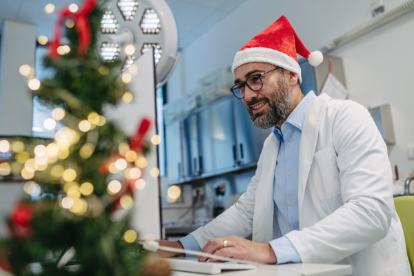 Portrait of handsome doctor in emergency room, consulting room decorated for Christmas. Mature doctor with Santa hat working a Christmas shift in the hospital, can't be with his family during Christmas Day and Christmas Eve.