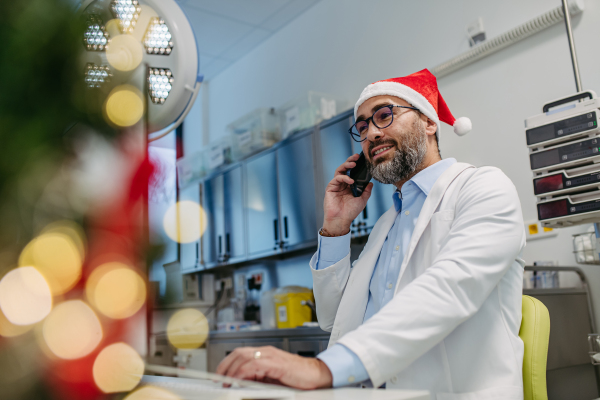 Portrait of doctor phone calling in emergency room, consulting room decorated for Christmas. Mature doctor with Santa hat working a Christmas shift in the hospital, can't be with his family during Christmas Day and Christmas Eve.