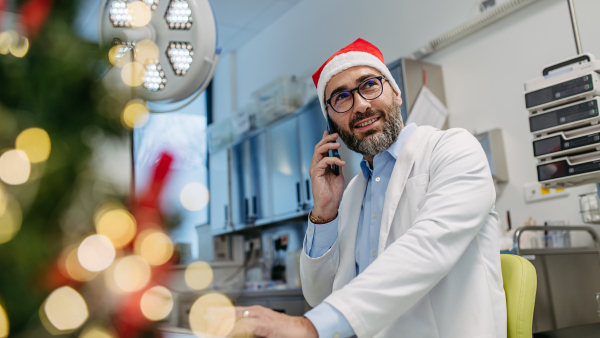 Portrait of doctor phone calling in emergency room, consulting room decorated for Christmas. Mature doctor with Santa hat working a Christmas shift in the hospital, can't be with his family during Christmas Day and Christmas Eve.