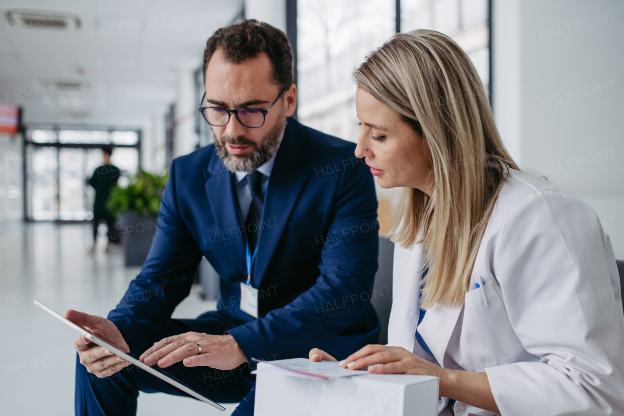 Portrait of pharmaceutical sales representative talking with doctor in medical building. Ambitious male sales representative in suit presenting new medication on tablet.