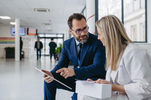 Portrait of pharmaceutical sales representative talking with doctor in medical building, clinic lobby. Ambitious male sales representative in suit presenting new medication, drugs, showing data on tablet.