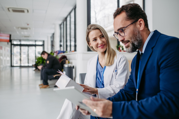 Portrait of pharmaceutical sales representative talking with doctor in medical building. Ambitious male sales representative in suit presenting new medication on tablet.