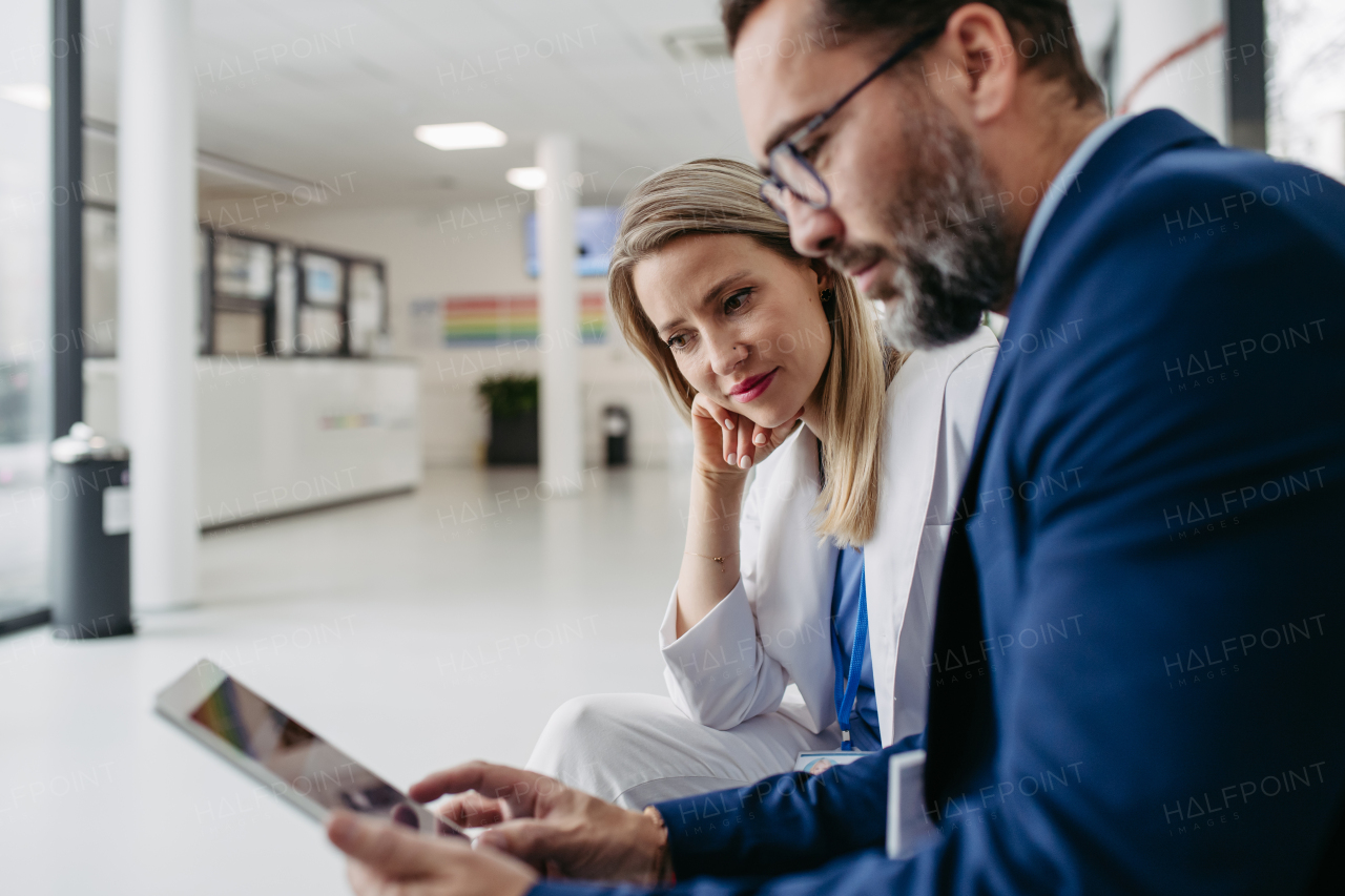 Portrait of pharmaceutical sales representative talking with doctor in medical building, clinic lobby. Ambitious male sales representative in suit presenting new medication, drugs, showing data on tablet.