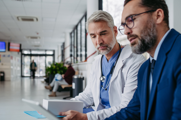 Pharmaceutical sales representative talking with doctor in medical building, presenting new medication on tablet. Hospital director or manager in modern clinic with surgeon.