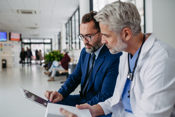 Pharmaceutical sales representative talking with doctor in medical building, presenting new medication on tablet. Hospital director or manager in modern clinic with surgeon.