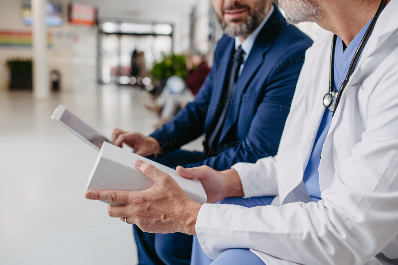 Portrait of pharmaceutical sales representative standing in hospital corridor. Hospital director, manager of private medical clinic in hospital lobby. Doctor sitting next to him.