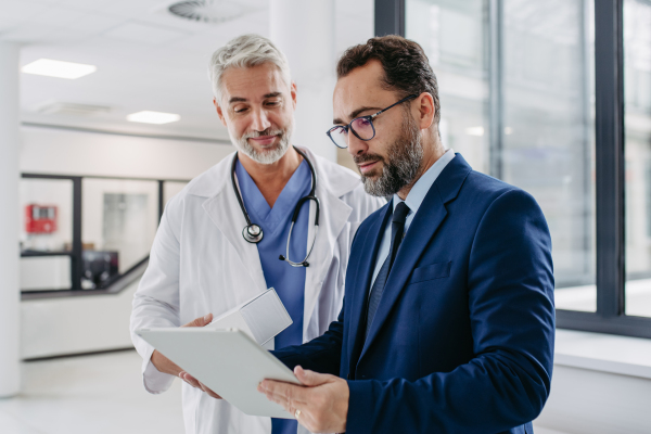 Pharmaceutical sales representative talking with doctor in medical building, presenting new medication on tablet. Hospital director or manager in modern clinic with surgeon.