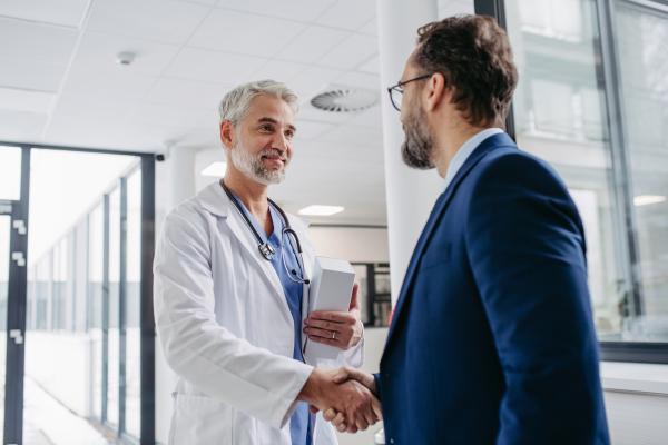 Portrait of pharmaceutical sales representative standing in doctor office, shaking hands. Hospital director, manager of private medical clinic greeting new doctor. Generous donor, giving donation to hospital.