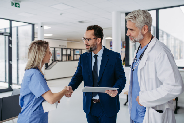 Pharmaceutical sales representative talking with doctors in medical building, presenting new product on tablet. Hospital director, manager in private medical clinic handshaking with doctors.