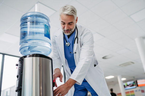 Handsome doctor is taking a break during work shift at hospital, drinking water from water dispenser in hospital lobby.