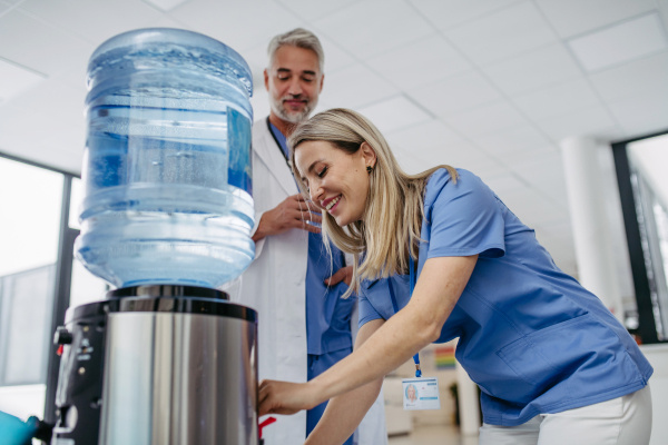 Handsome doctor and nurse taking a break during work shift at hospital, drinking water from water dispenser in hospital lobby, talking.