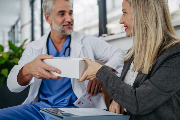 Female pharmaceutical sales representative presenting new medication to doctor in medical building, holding box with medication, drugs samples.