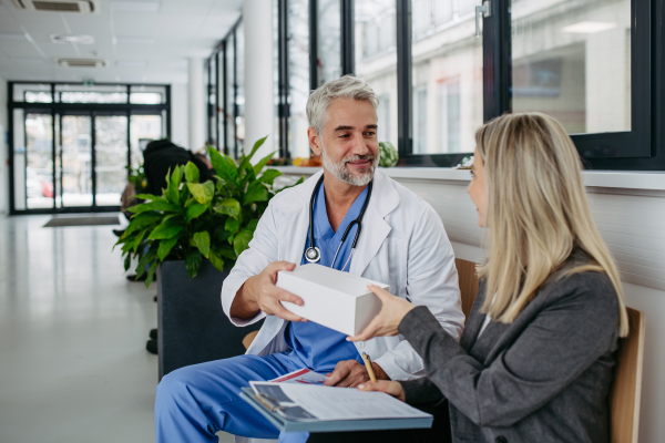 Female pharmaceutical sales representative presenting new medication to doctor in medical building, holding box with medication, drugs samples.