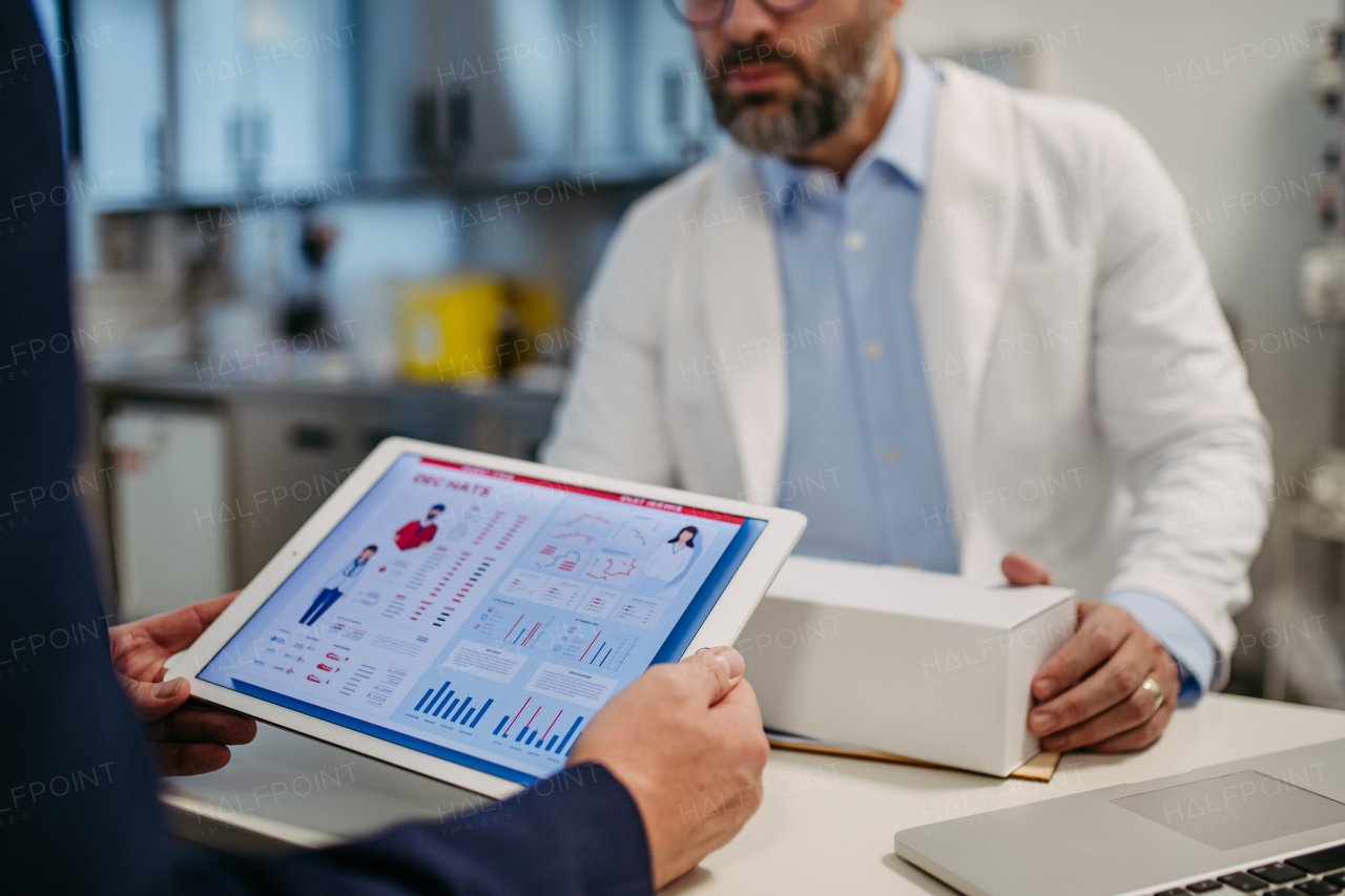 Pharmaceutical sales representative presenting new medication to doctor in medical building, holding tablet with presentation, graphs and statistics, explaining important informations.