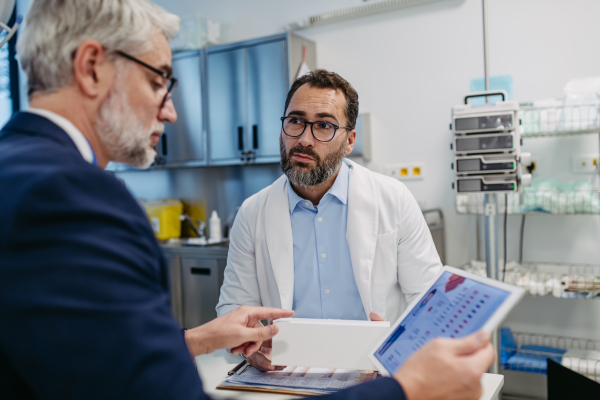 Pharmaceutical sales representative presenting new medication to doctor in medical building, holding tablet with presentation, graphs and statistics, explaining important informations.