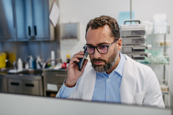 Portrait of ER doctor in hospital working in emergency room. Healthcare worker in white coat making call and typing on medical computer in emergency room.