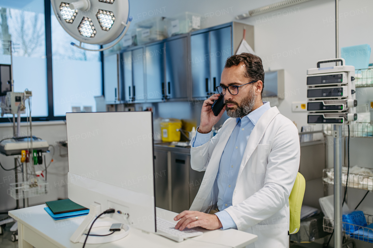 Portrait of ER doctor in hospital working in emergency room. Healthcare worker in white coat making call and typing on medical computer in emergency room.