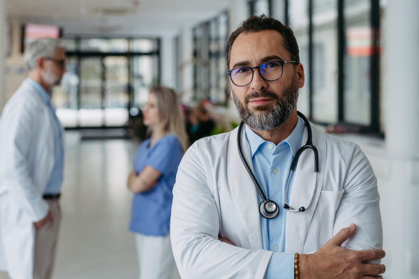 Portrait of confident mature doctor standing in Hospital corridor. Handsome doctor wearing white coat, scrubs, stethoscope around neck standing in modern private clinic, looking at camera.