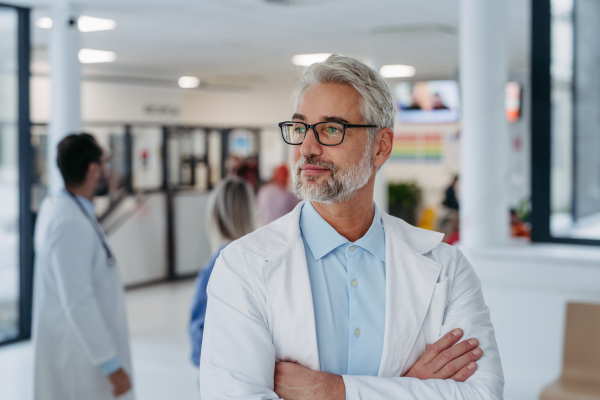 Portrait of confident mature doctor standing in Hospital corridor. Handsome doctor with gray hair wearing white coat, scrubs, stethoscope around neck standing in modern private clinic, looking at camera.