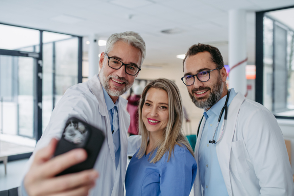 Portrait of doctors and nurse standing in hospital corridor. Healthcare workers in modern private clinic, taking selfie and smiling.