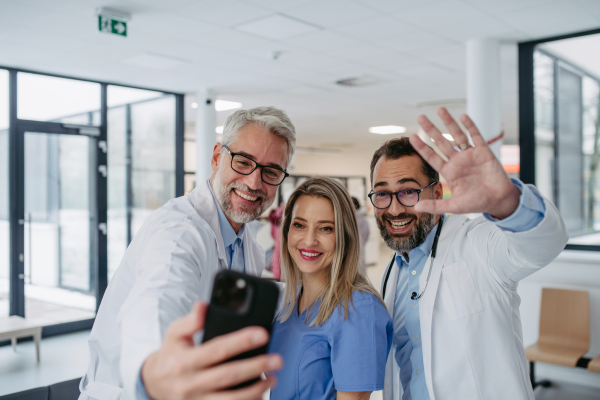 Portrait of doctors and nurse standing in hospital corridor. Healthcare workers in modern private clinic, taking selfie and smiling.