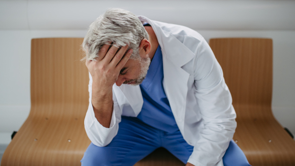 Low angle shot of frustrated, exhausted doctor sitting in hospital corridor. Concept of burnout syndrome among doctors, health care workers.