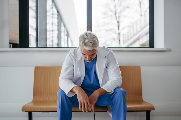 Frustrated, exhausted doctor sitting on bench in hospital corridor. Concept of burnout syndrome among doctors, health care workers.